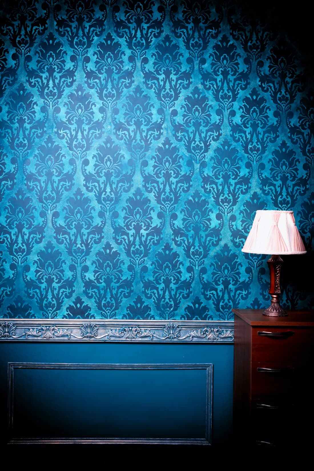 Deep teal damask wallpaper behind a lamp and painted dresser